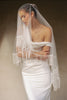 A woman wearing a white strapless wedding dress and a long sheer bridal veil with fringe trim, posed against a warm beige studio background.
