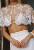 A close-up of a model wearing a white lace bustier crop top over a white bralette and a white high-waisted skirt. The lace top features intricate floral patterns and a keyhole neckline.