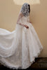 A woman wearing a voluminous white lace wedding gown with a long sheer veil, photographed from behind against a plain beige wall.