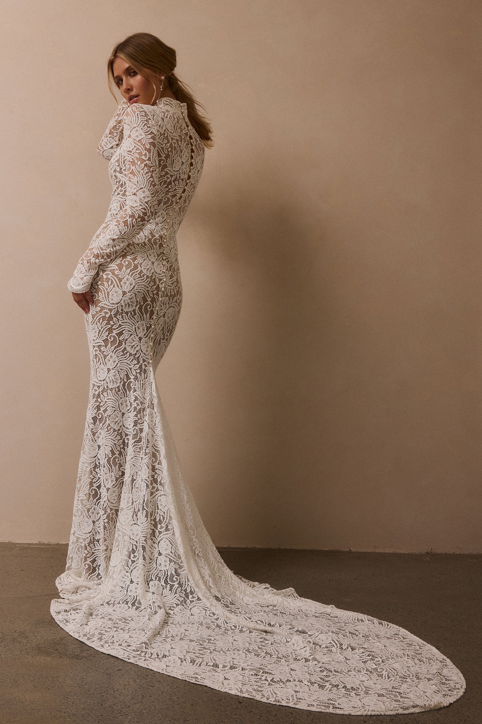 A woman wearing a fitted white lace wedding gown with long sleeves and a dramatic train, photographed in a studio with a warm beige wall and dark floor_XS_