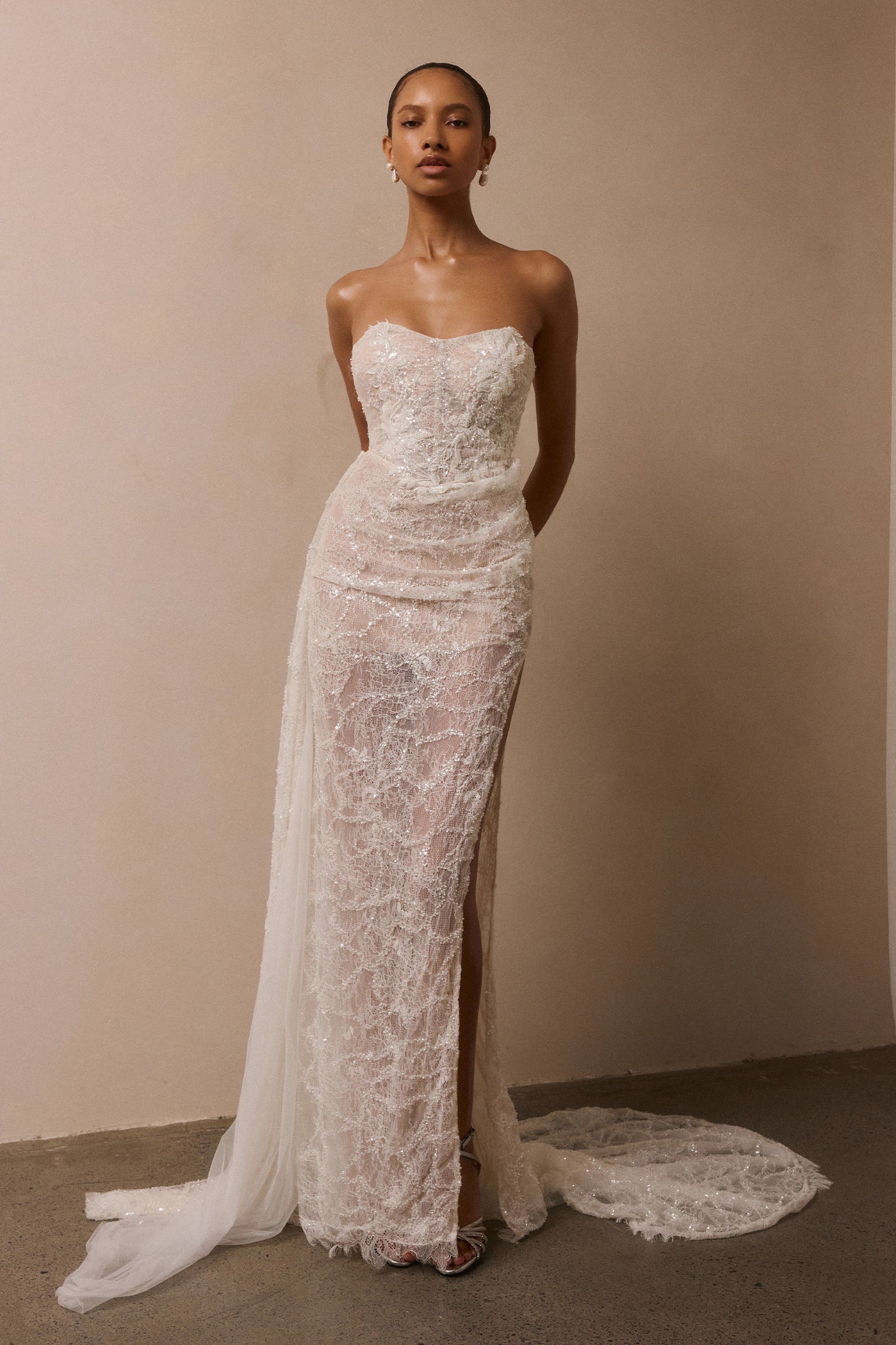 A woman modeling a strapless white lace wedding gown with a fitted bodice, thigh-high slit, and long sheer train against a plain beige wall_XS_