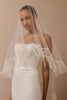 A woman wearing a long sheer white bridal veil with beaded fringe draped over her shoulders and a white strapless gown against a warm beige studio background.