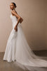 A woman modeling a sleeveless white wedding gown with a fitted bodice, full skirt, and long sheer tulle train against  a plain beige studio wall_XS_