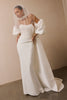 A woman wearing a strapless ivory wedding gown with dramatic puffed sleeves, a sheer veil, and a long train, standing against a plain beige wall.