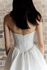 A woman shown from behind wearing a strapless white dress with a fitted textured bodice and full skirt, standing against a light brick wall.