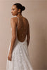 A woman wearing a white lace bridal gown with a dramatic open back, layered with delicate silver chain necklaces, photographed against a solid beige studio background.