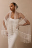 A woman wearing a white strapless gown and a sheer white bridal veil with long fringe, posed against a warm beige studio background.