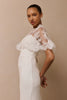 A woman modeling a white wedding dress with sheer lace cape against a plain beige studio background.