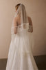 A woman seen from behind wearing a white wedding dress with fringe detailing and a long sheer bridal veil, standing against a plain beige wall.