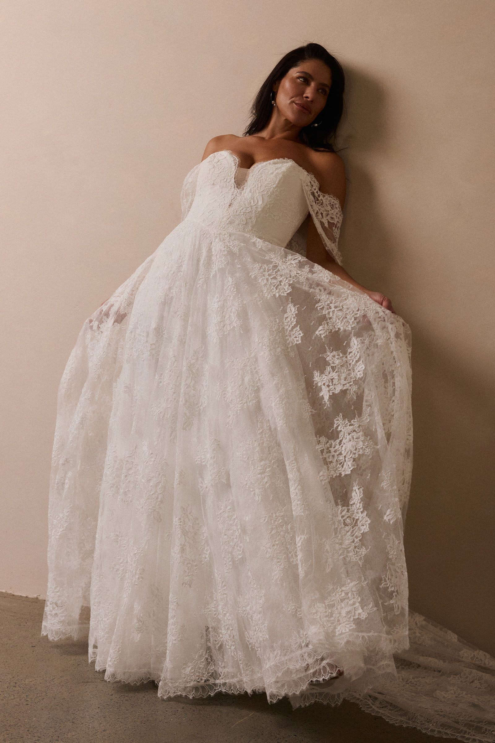 A woman wearing a white off-the-shoulder lace wedding gown with a deep V neckline and a full layered skirt_L_