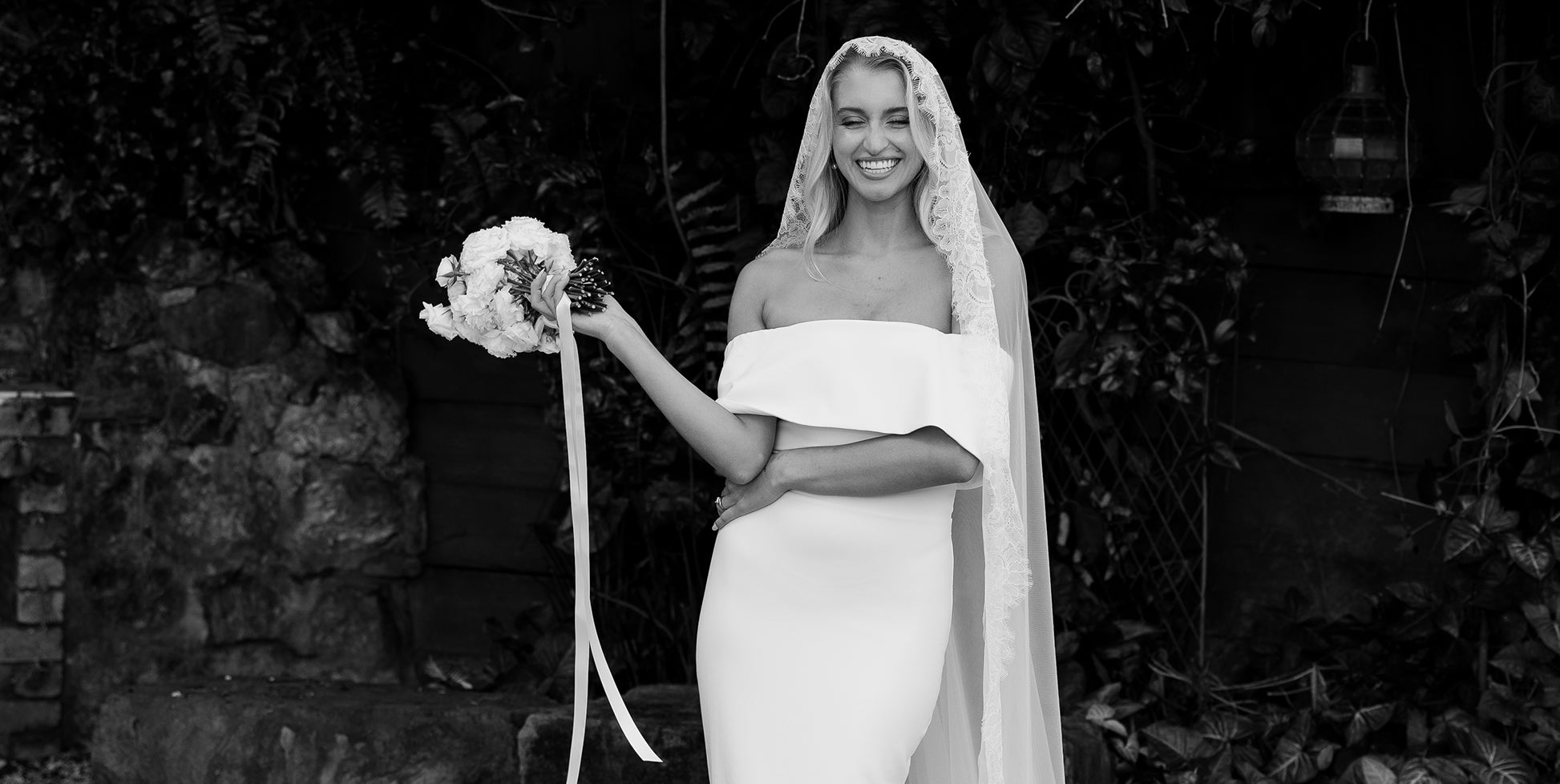 A bride wearing an off the shoulder crepe wedding dress holding flowers.